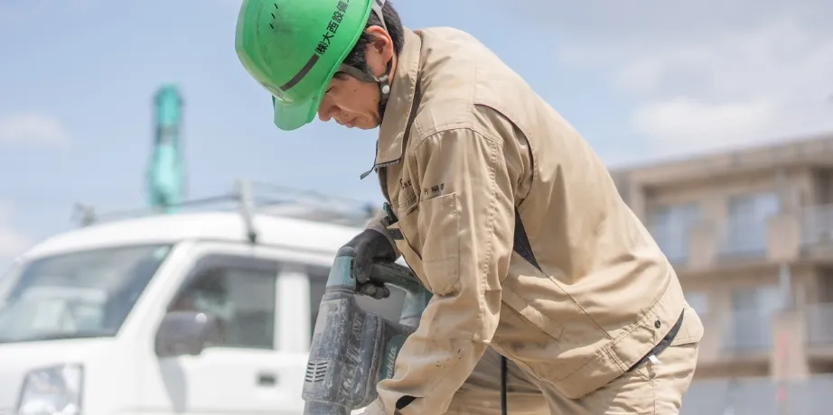 緑色のヘルメットと作業服を着た男性が、電動工具を使用して地面の作業を行っている様子