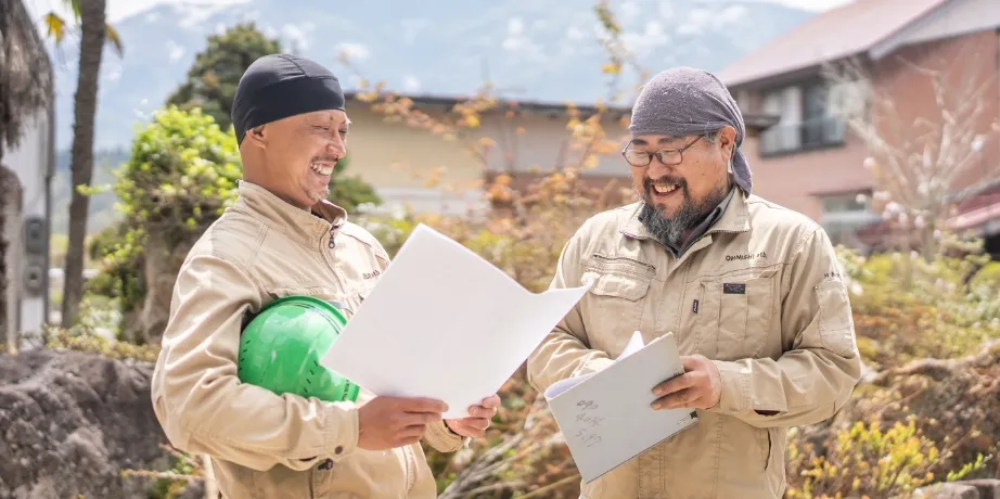 作業服を着た2人の男性が屋外で書類を見ながら笑顔で話し合っている様子