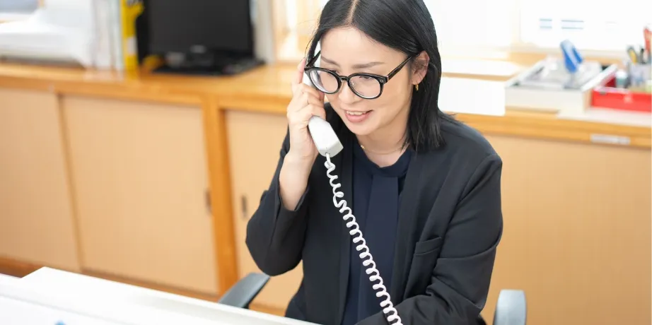 黒髪で眼鏡をかけた女性が、明るいオフィス内で固定電話を使って応対している様子
