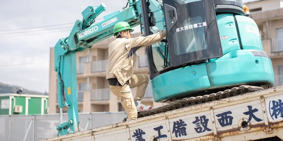 緑色のヘルメットを着用した作業員が、トラック荷台に載せたショベルカーに乗り込もうとしている様子