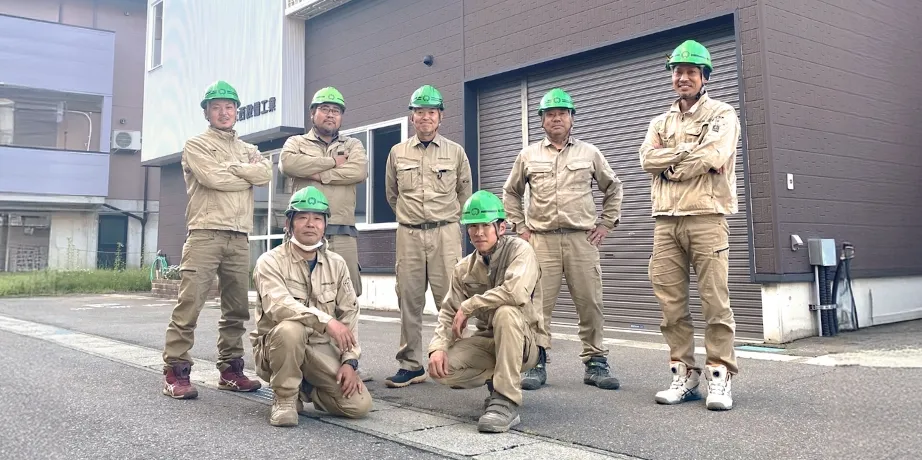 作業服と緑色のヘルメットを着用した7人の作業員が、建物前でポーズをとって集合写真を撮っている様子