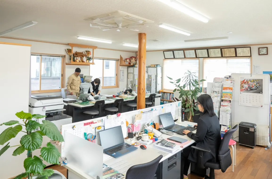 複数の人がデスクでパソコン作業や打ち合わせを行っているオフィス内の様子