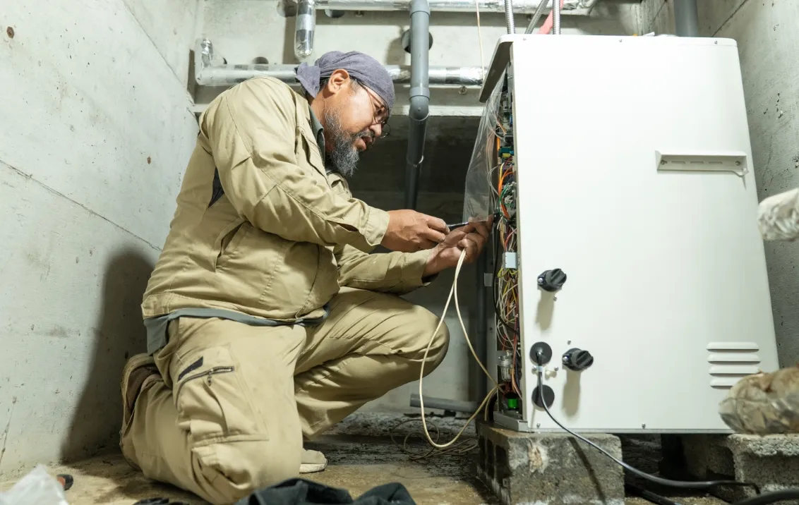 作業着を着た男性が、機械内部の配線を確認しながら電気系統の作業を行っている