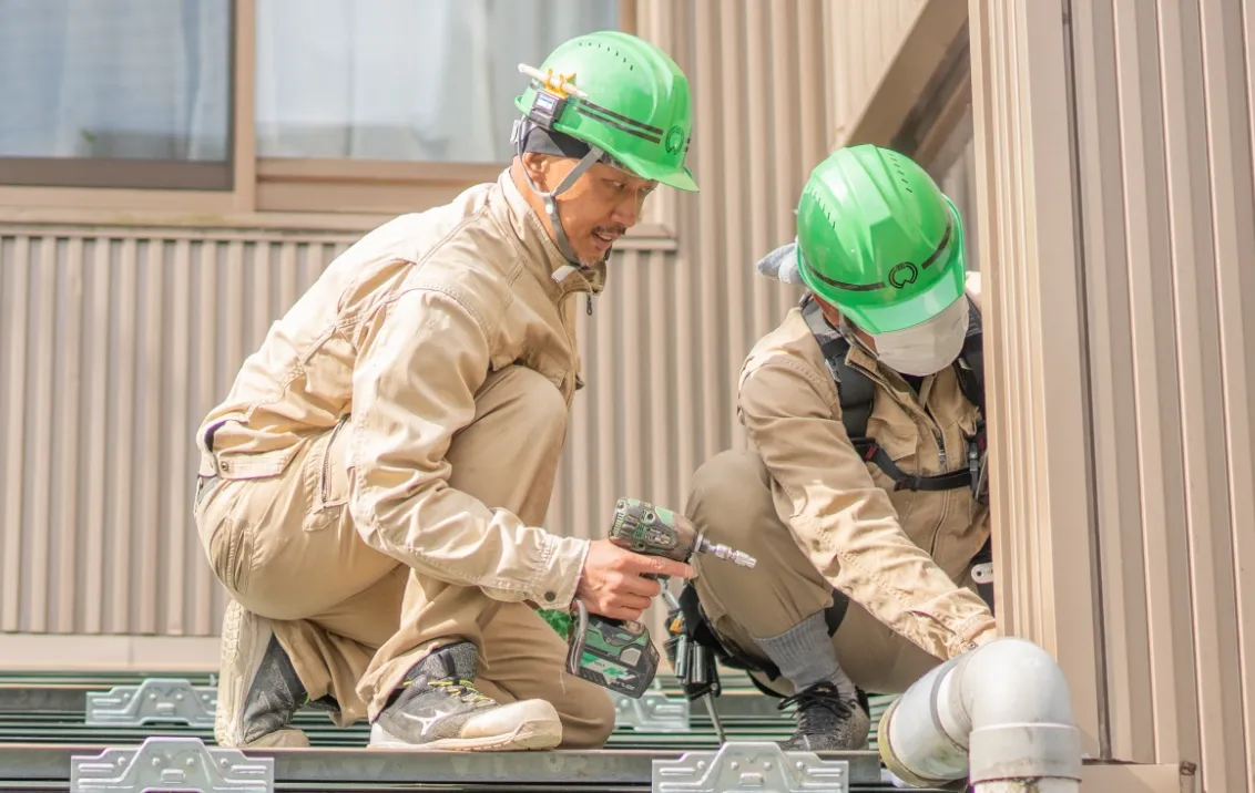 緑色のヘルメットを着用した2人の作業員が、屋外で配管や設備の取り付け作業をしている