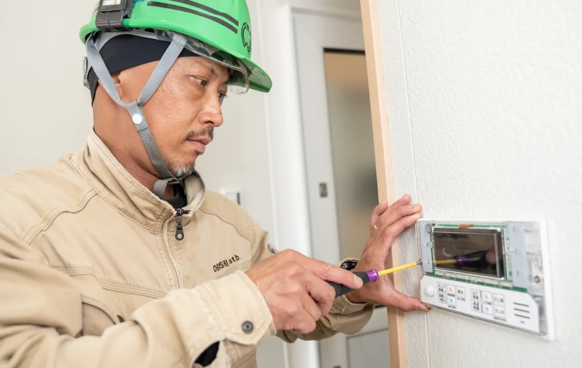 緑色のヘルメットをかぶった作業員が、壁に取り付けられた機器を工具で調整している