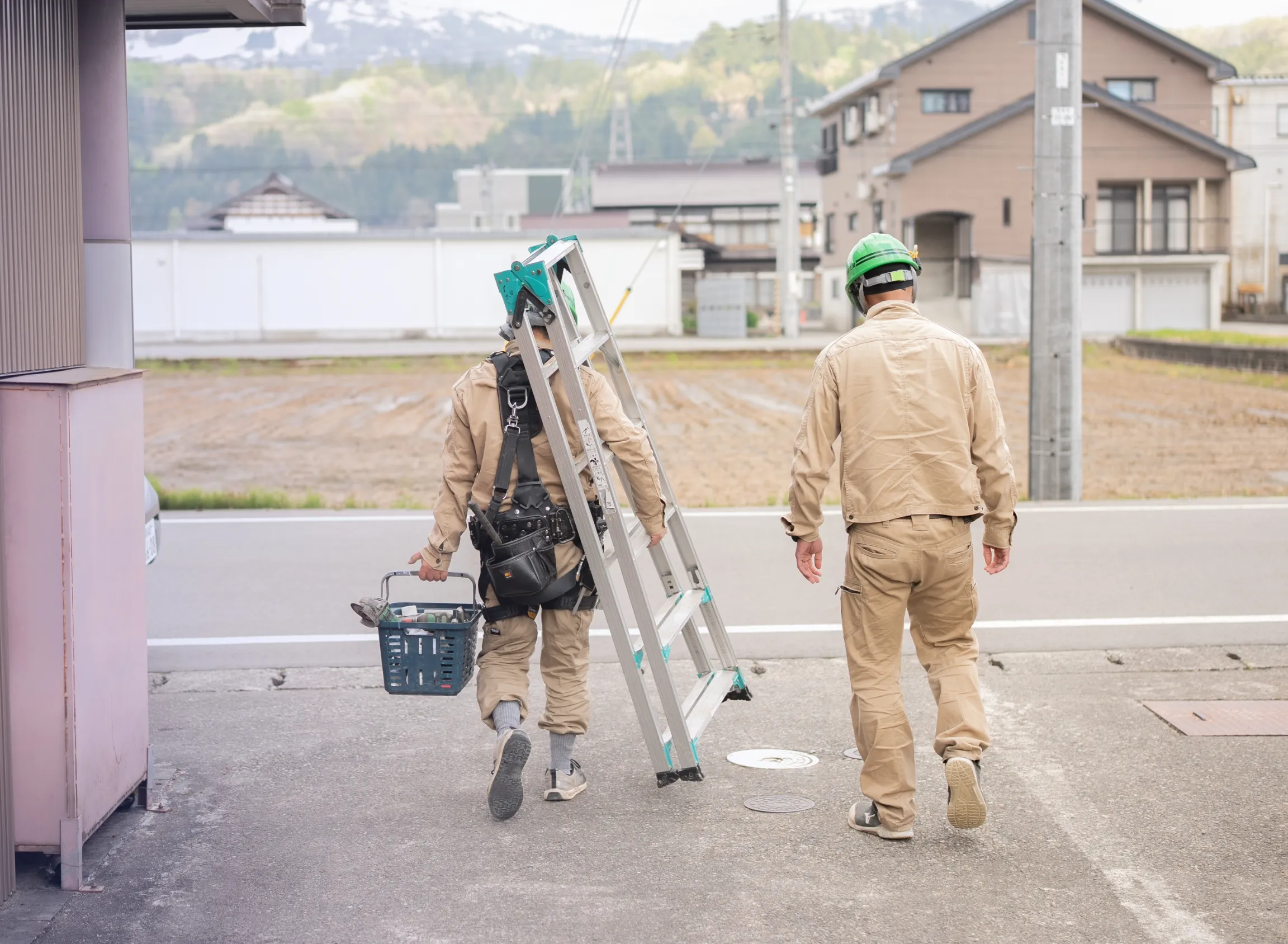 作業員2人がハシゴや工具を持って現場に向かって歩く後ろ姿