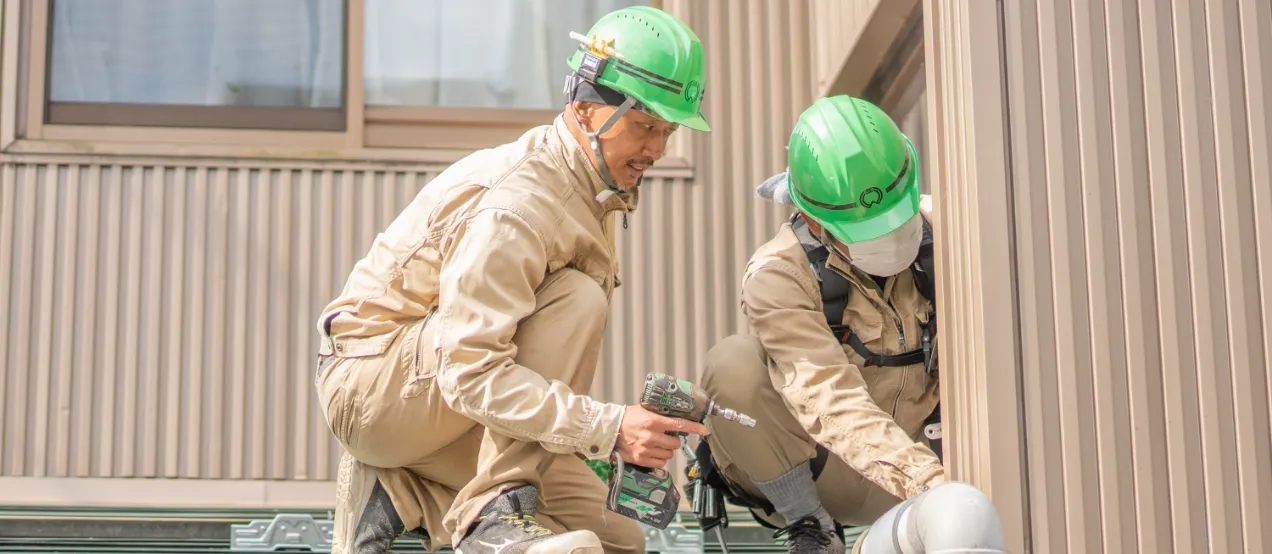 緑色のヘルメットと作業服を着た2人の作業員が、建物の外壁で設備の取り付け作業をしている