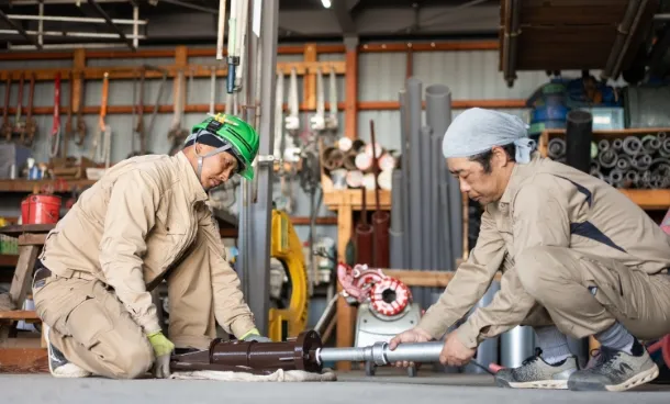 作業服を着た2人の作業員が、工場や作業場内で金属製のパイプを機械に取り付けている様子