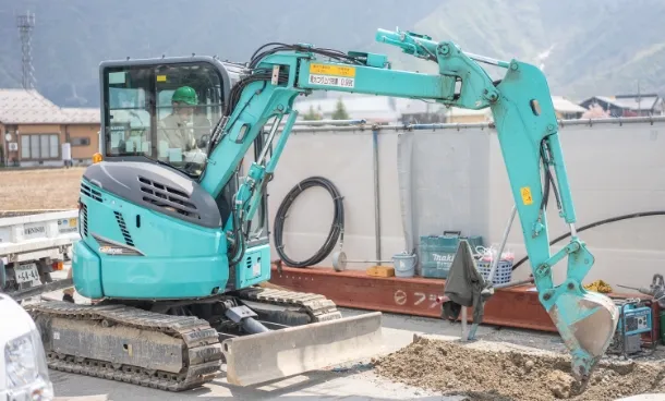 建設現場で、緑色のヘルメットをかぶった作業員が小型ショベルカーを操作している様子